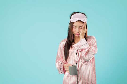 Sleepy Asian woman in pajamas with mask is holding a cup, wanting sleep and rest at home, feeling tired and dissatisfied, needing relaxing night nap to calm her mood, isolated studio blue background
