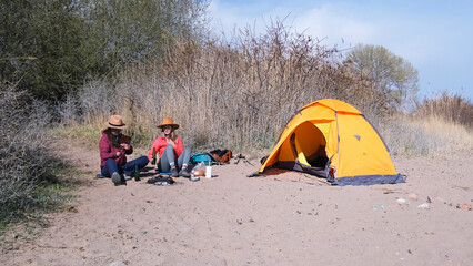 Two tourists are drinking on the beach next to a tent. Two women are having fun outdoors with beer bottles in their hands