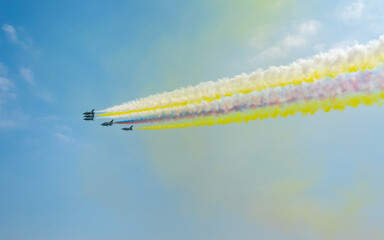 Multiple planes fly in formation, leaving trails of white, yellow, and red smoke against a blue sky.