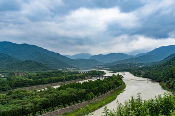 Dujiangyan river landscape Sichuang China shows winding waterways surrounded by lush green mountains under a cloudy sky, with a long structure spanning the water.