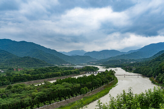 Dujiangyan river landscape Sichuang China shows winding waterways surrounded by lush green mountains under a cloudy sky, with a long structure spanning the water.