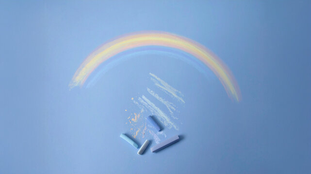 A vibrant, hand-drawn chalk rainbow arcs gracefully over pastel chalk sticks and playful scribbles on a serene blue background. This artistic composition evokes childhood wonder, creativity, and the j