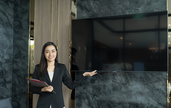 Beautiful Asian Business Woman in black suit pointing to present screen Wall TV Showing Digital Entrepreneur presenting a project on empty blank screen TV mock up presentation.