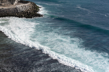 A powerful ocean swell forms a perfect wave, breaking with white foam over a dark reef near a cliffside breakwater in Bali, Indonesia.