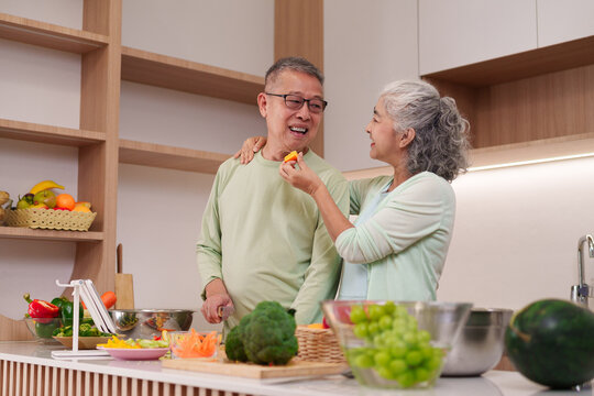 Asian older adult couple happily preparing healthy fresh food home kitchen sharing fruit lovingly cooking wholesome meal together modern vibrant kitchen - Powered by Adobe