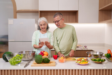 Asian senior couple cooking healthy fresh food at home kitchen using digital tablet for recipe guidance joyful elderly pair preparing nutritious meal together domestic setting