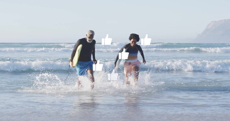 Running senior man and woman carrying yellow surfboard from surf at shoreline, wearing rash guards