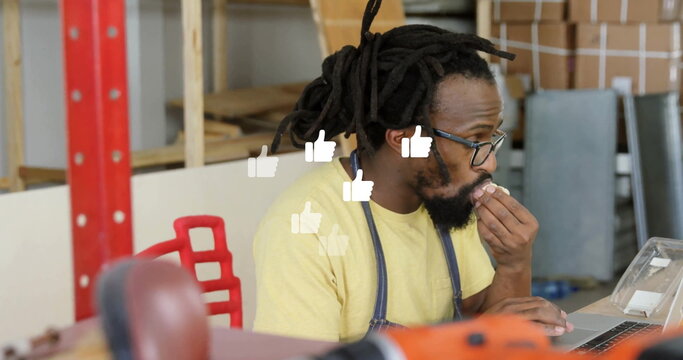 Typing craftsman with dreadlocks wearing denim apron eating snack at workbench, laptop, hand truck - Powered by Adobe