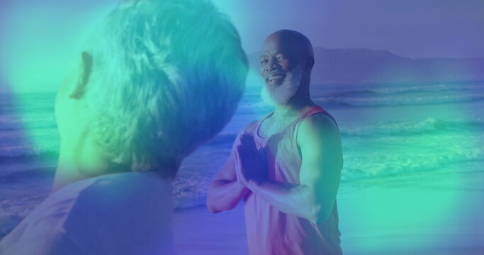 Smiling African American man wearing coral top pressing hands in yoga prayer at beach, with friend