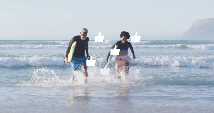 Running senior man and woman carrying yellow surfboard from surf at shoreline, wearing rash guards