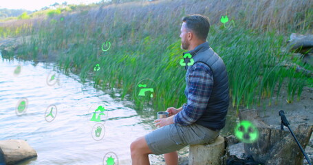 Gazing man sitting on stump holding stainless steel mug at lakeshore, with green icons, copy space