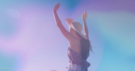 Raising arms woman wearing straw sun hat, floral sleeveless top and denim shorts under gradient sky