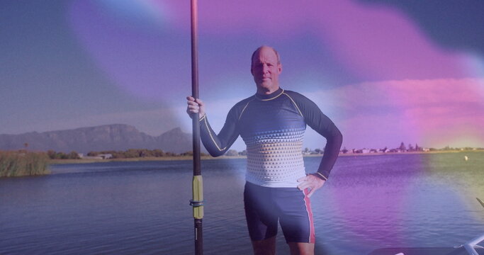 Standing senior male athlete in athletic wear holding rowing oar at calm lake shore, with boat - Powered by Adobe