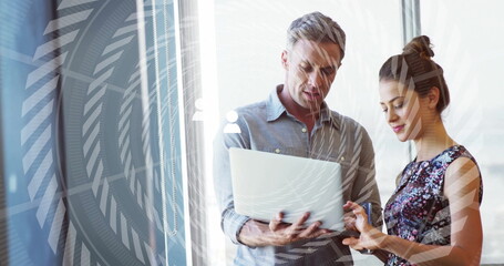 Colleagues in formal attire reviewing silver laptop content in office tower, with biometric overlay