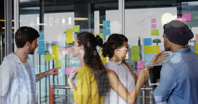 Collaborating team of four reviewing ideas on glass partition in open office, with sticky notes