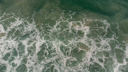 Aerial view of ocean waves crashing with white foam on green sea surface, creating natural texture and abstract pattern, perfect for backgrounds, nature concepts, and coastal design.
