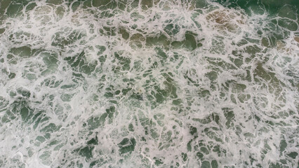 Aerial view of ocean waves crashing with white foam on green sea surface, creating natural texture and abstract pattern, perfect for backgrounds, nature concepts, and coastal design.