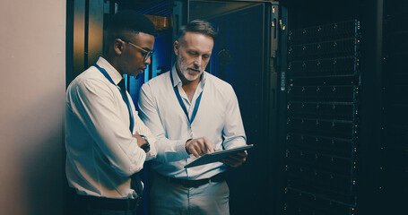 Men, tablet and talk in server room with inspection, review and system maintenance report with mentor. People, tech and discussion with application, feedback and programming with cybersecurity team