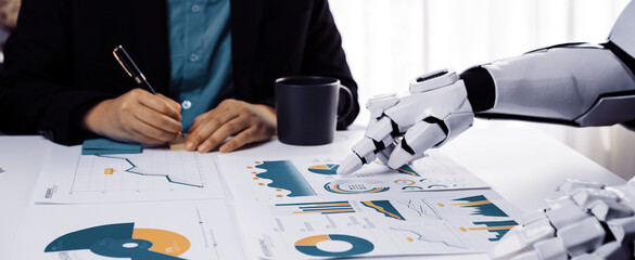 A human and a robotic hand engage in a collaborative business meeting, analyzing detailed financial charts in a modern office, highlighting innovation and technology.Noogenesis