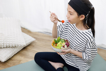 A healthy Asian girl in casual clothes with a headband sitting on the floor, eating a delicious bowl of salad for breakfast. She's focused on getting the best nutrition.
