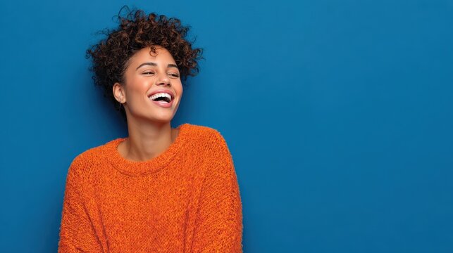 happy emotions concept positive dark skinned beautiful young woman laughs poisitively looks aside with carefree face expression wears casual orange sweater isolated over blue studio background no log