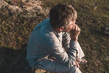 man rests on rock, lost in thought as he gazes into distance. late afternoon sun casts soft shadows across grassy field surrounding him, creating serene atmosphere. close up.