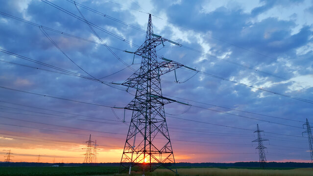 Power lines and pylons silhouetted against a vibrant sunset sky, blending industrial strength with natural beauty, captured in an establishing shot. - Powered by Adobe