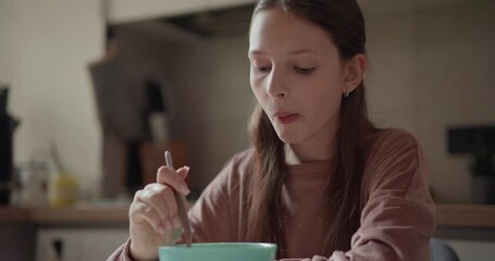 Close up of teenage girl eating cereal with milk from blue bowl during her breakfast in kitchen in the morning. Morning breakfast and kitchen routine