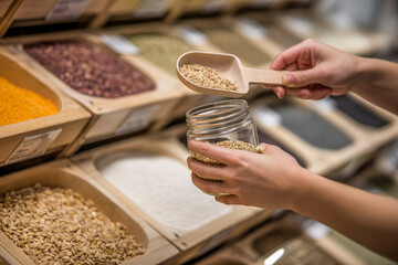 Woman's hands filling small glass jar with grains using wooden scoop at bulk store. Concept of zero-waste and sustainable shopping