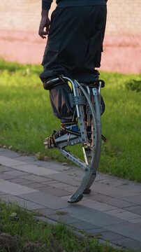 Person walking on stilts wearing gray hoodie and pants on paved road surrounded by green grass and trees, showcasing balance, agility, and dynamic motion, with an outdoor setting