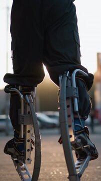 Back view of person walking on spring stilts during sunset wearing gray hoodie and pants. Background features blurred trees, suburban street, and warm sunlight creating soft shadows