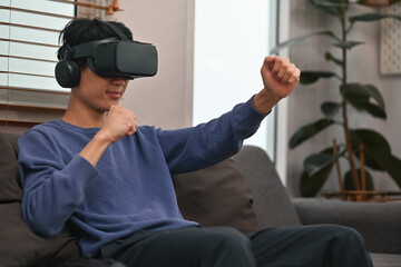 A Young Asian man simulates action while using VR headset on couch, relaxed and entertained