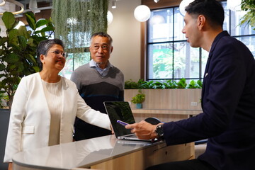 Senior male and female tourists dragging luggage arrive at the hotel. Standing at the counter, a male receptionist welcomes them, registers them into the room, and provides accommodation information.