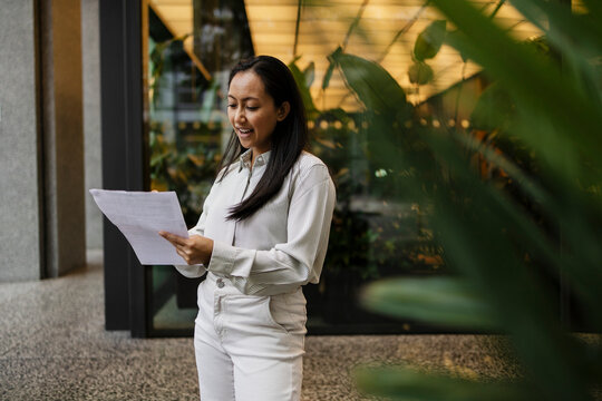 Businesswoman reading document outdoors near modern office building