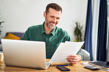 Smiling man working from home using laptop holding documents