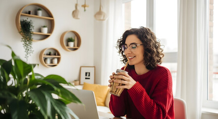 Happy woman in red sweater enjoying coffee while working on laptop in bright modern home office with decorative shelves and plants