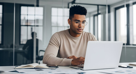 Obraz premium Young man working on a laptop in a modern office space with large windows and natural light for business and technology themes