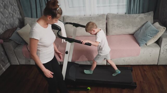 Young boy in white shirt and green socks jumps playfully on treadmill while woman in white top and black pants tries to guide him, highlighting contrast between instruction and playful behavior