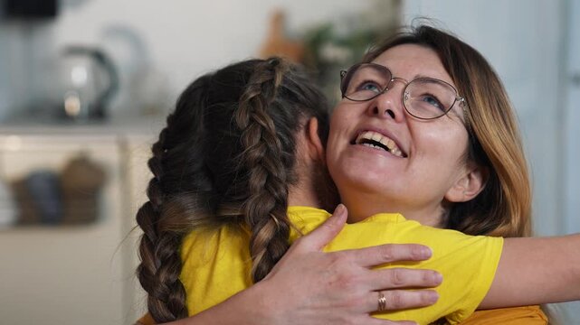 Mother hugs daughter warmly in kitchen, smiling and embracing child with braided hair and yellow shirt, family love and affectionate parent bond, mother and daughter hug and smile in cozy home