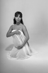 Fototapeta premium Black and white portrait of a young pregnant woman covering her body with a translucent white cloth and sitting on her knees in full length