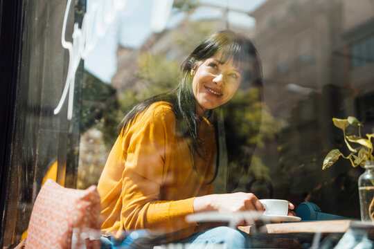Happy woman with coffee cup sitting in cafe seen through glass