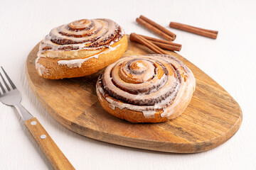 Two homemade freshly baked cinnamon rolls bun or swirl decorated with sugar icing served on wooden cutting board with spice sticks on white table with fork for breakfast as sweet dessert pastry
