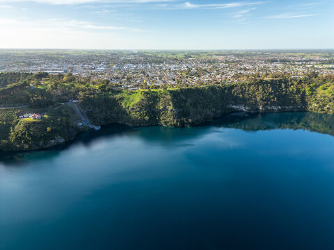 Aerial view of the Blue Lake's striking cobalt waters contrast with the verdant, cliffside foliage, shadowed by the distant city skyline, Mount Gambier, South Australia, Australia.