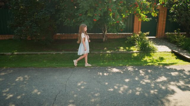 Fair skin girl with long blonde hair walks alone by roadside in residential area, bordered by green lawn, brick fence, and lush tree casting soft shadows on sunny day