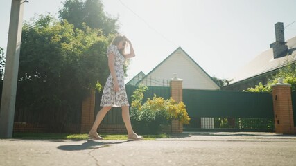 Young woman in floral dress walking calmly under intense midday sunlight, passing by green fence and utility pole, with shadows cast on road and suburban house in background