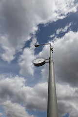 Low Angle View Of A Street Clock And Lamppost Against A Dramatic Cloudy Sky. Urban Detail And Time Concept. Vertical Shot.