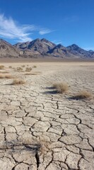 Cracked earth spans a vast, arid landscape leading to hazy, brown mountains under a bright blue sky with wispy clouds