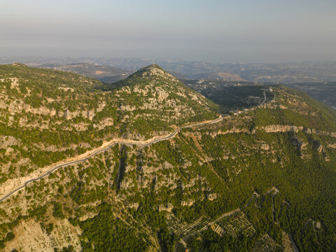 Aerial view of a winding road slicing through verdant mountains, dotted with limestone rocks under a soft, hazy sky, Douma, North Governorate, Lebanon.