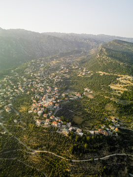 Aerial view of terracotta rooftops nestled amidst verdant trees and rugged terrain, bathed in the warm glow of the setting sun, Douma, North Governorate, Lebanon.