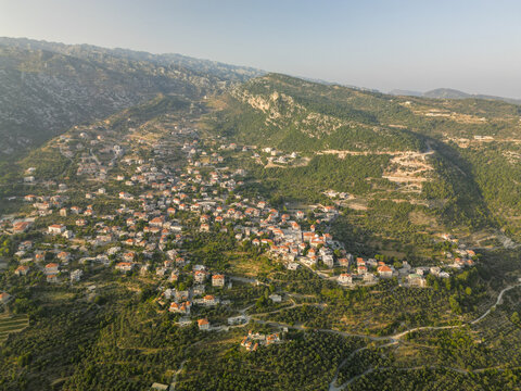 Aerial view of a cluster of terracotta-roofed houses nestled amidst the green hills and rugged terrain, illuminated by the soft glow of the setting sun, Douma, North Governorate, Lebanon.